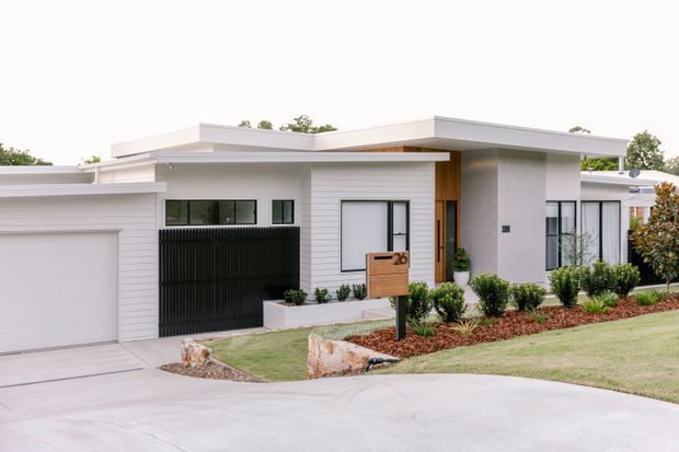 Street-facing view of a contemporary single-storey residence with a low-pitched flat roof, crisp white horizontal cladding and rendered walls, dark vertical slatted screening, and warm timber detailing at the recessed entry. The home includes an attached single garage, landscaped front garden beds, a concrete driveway, and a timber letterbox.