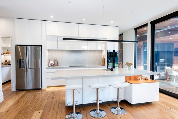 Bright contemporary kitchen with extensive glossy white cabinetry, a large pale stone island with three bar stools, stainless-steel refrigerator, integrated appliances, timber flooring, and a long linear pendant light. Full-height black-framed glazing opens visually to an outdoor deck or terrace.