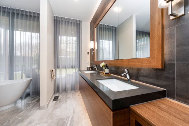 Contemporary bathroom featuring a freestanding white bathtub beside tall sheer-curtained windows, a long timber vanity with a white integrated basin, dark stone benchtop, large framed mirror, and dark tiled walls. The room has a bright, refined atmosphere with warm timber accents and natural light.