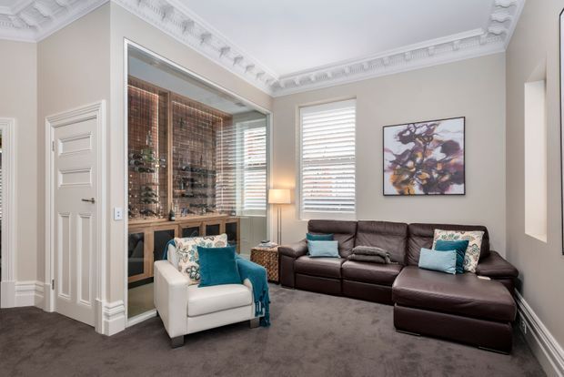 A refined living room with ornate decorative plaster cornices and wall mouldings, neutral painted walls, plush grey carpet, and a dark brown leather sectional sofa. A white upholstered accent chair with turquoise cushions sits beside a glass-fronted built-in wine display cabinet, while plantation shutters, a floor lamp, and abstract artwork complete the room.