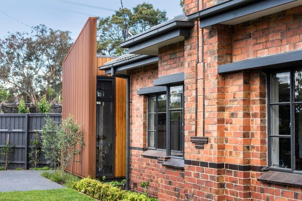 Exterior view of a contemporary brick residential building with black-framed windows, projecting dark metal eaves, and a vertical timber-clad entry volume. The facade combines exposed red brick with warm timber and dark detailing, opening onto a landscaped garden and paved path.