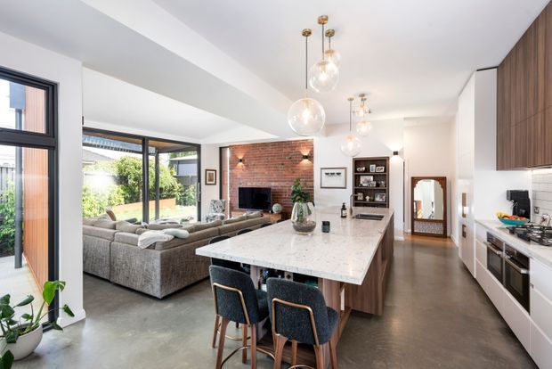 Bright open-plan kitchen and living area with a large marble-look island, dark upholstered bar stools, white cabinetry, timber upper cabinets, and a tiled backsplash. The adjacent living room includes a large sectional sofa and a brick fireplace feature wall, with extensive black-framed glazing opening toward a garden. Globe pendant lights hang over the island, and the space has polished contemporary detailing with warm timber accents.