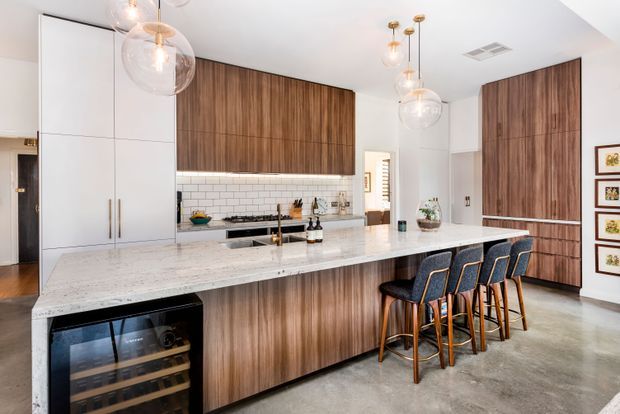 A bright, contemporary open-plan kitchen dominated by a large white stone island with integrated seating for four. The kitchen features extensive vertical-grain timber cabinetry, tall white pantry cabinetry, a white subway-tile splashback, integrated appliances, polished metal fixtures, and glass globe pendant lights. The polished concrete floor and restrained palette create a refined, warm, high-end interior.