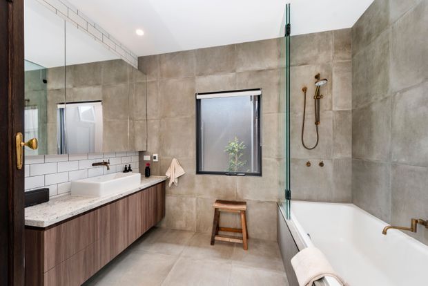 A contemporary bathroom with a long floating timber vanity, white vessel basin, pale stone benchtop, large wall mirror, white subway-tile backsplash, and grey-beige large-format tiled walls and floor. A freestanding bathtub sits beside a clear glass shower screen, with brass fixtures and a small timber stool adding warmth. The room is bright, clean, and refined.