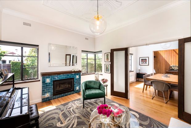 Bright traditional living room with white walls, ornate pressed ceiling detailing and decorative cornices, black-framed windows, a teal tiled fireplace with timber mantel, green velvet armchair, patterned rug, piano, and glass coffee table. Open dark timber-framed doors connect to a warm timber-floored dining room with a large wooden table and a vertical timber feature wall.