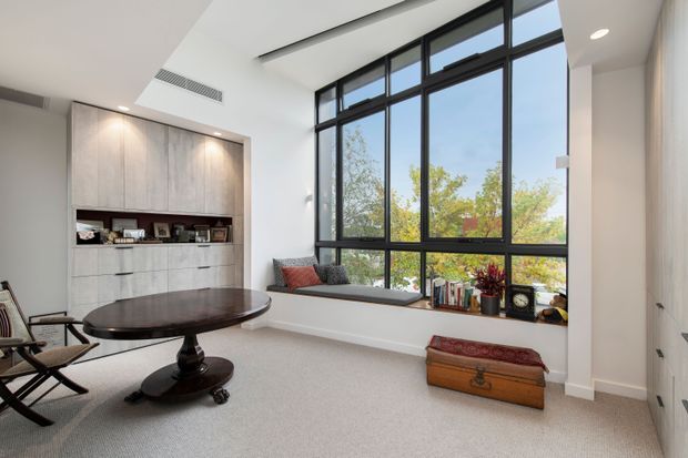 Bright, contemporary sitting room with a large black-framed floor-to-ceiling window, a built-in light timber display and storage wall, window seat, round dark wood pedestal table, and lounge chairs. The room has a high ceiling with recessed lighting and a neutral carpeted finish, with trees visible outside.
