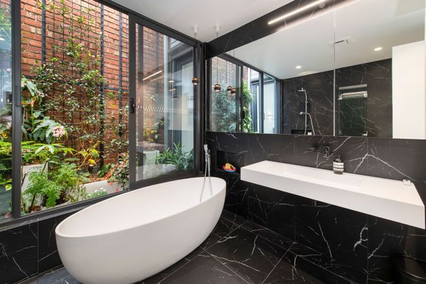 Contemporary bathroom with a freestanding white oval bathtub beside floor-to-ceiling black-framed glazing. The glazing overlooks a lush enclosed courtyard and exposed red-brick wall. Black marble-look wall and floor surfaces with prominent white veining contrast with a floating white vanity and integrated basin. A frameless shower and large mirror are visible, along with recessed ceiling lights and a linear pendant.