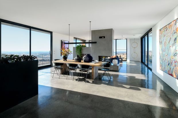 A bright, open-plan contemporary dining and living area with floor-to-ceiling glazing, expansive views, polished concrete floors, and a long timber dining table with black wire-frame chairs. A central concrete fireplace or chimney volume anchors the space, while large abstract artwork and indoor planting add colour and texture.