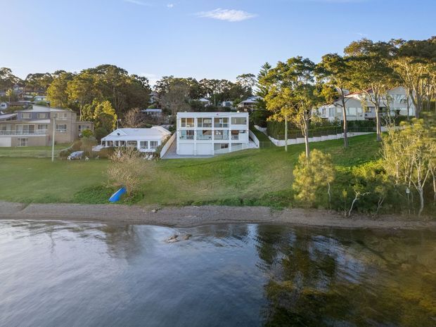 Aerial exterior view across a calm waterfront toward several residences on a gently sloping, landscaped shoreline. The central modern white multi-level house has broad glazing and balconies, flanked by neighbouring homes and mature trees. A narrow pebbly beach and reflective water occupy the foreground.