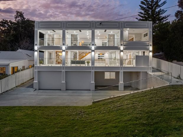 Rear exterior view of a contemporary multi-storey residential house at dusk, with a highly glazed two-level facade, grey rendered walls, illuminated interiors, balconies or terraces, and a sloped landscaped site with access stairs and driveway.