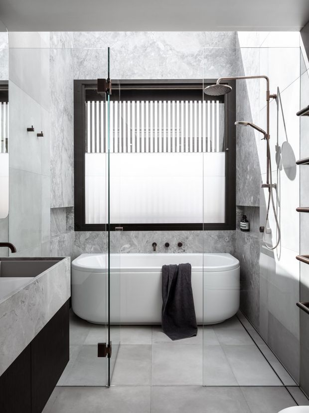 Contemporary bathroom featuring a freestanding white bathtub beneath a large black-framed window, enclosed by a clear glass shower screen and surrounded by pale grey marble-look wall and floor tiles. A dark timber vanity is partially visible on the left, with a wall-mounted shower system and heated towel rail on the right. The space is bright, clean and highly refined.