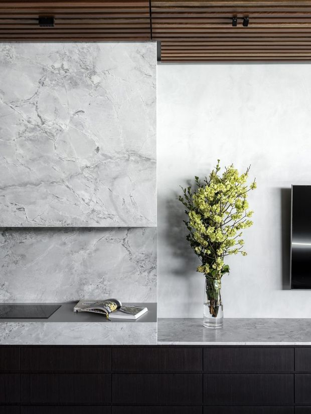 A contemporary living room media wall featuring dark built-in cabinetry, a grey veined stone surface, and a large wall-mounted television at right. The white plaster wall is paired with a warm timber slatted ceiling, while a glass vase with flowering branches sits on the stone console. A low-profile black fireplace or inset feature is visible at left.
