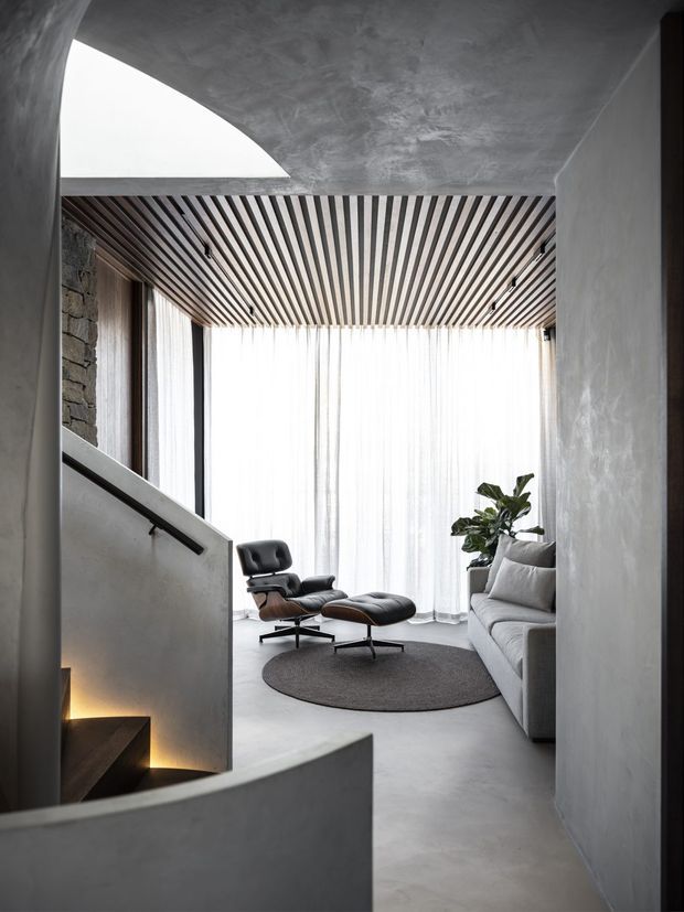 A refined, minimalist living room viewed from a sculptural stairway. The space features a grey plastered curved ceiling and walls, a linear timber slatted ceiling, floor-to-ceiling sheer curtains, a grey sofa, black leather lounge chair with matching ottoman, round dark rug, indoor plant, and polished concrete-style flooring. A rough stone wall is visible beside the glazing, while concealed stair lighting adds warmth.