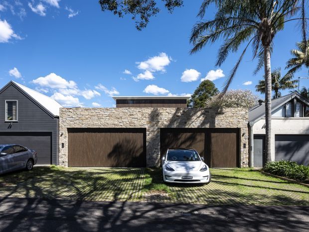 Street-facing view of a contemporary residential garage frontage with two wide timber garage doors set into a pale rough-stone wall. A dark-clad gabled house is visible on the left and a light-rendered gabled structure on the right, with a low rooftop volume behind the garage wall. The driveway uses grass-filled grid pavers, and mature palm trees and planting frame the sunny suburban scene.