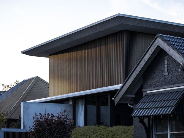 Exterior view of a contemporary multi-level residential building with a prominent broad flat roof over a tall upper volume clad in narrow vertical timber-like battens. Dark grey pitched-roof forms, corrugated roofing, textured masonry, deep eaves, and large glazed openings create a layered composition. Low hedging and shrubs partially screen the ground level.