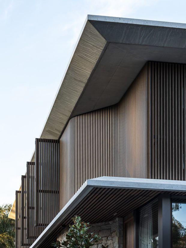 Close-up architectural exterior view of a contemporary multi-level building with bold projecting concrete roof and balcony slabs, extensive vertical dark timber or metal screening, and a textured stone wall at the lower level. The image emphasizes strong geometric lines, deep overhangs, layered balconies, and a sheltered entry or terrace area.