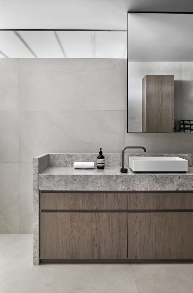 Contemporary minimalist bathroom vanity with a large rectangular mirror, wall-mounted timber-look cabinetry, a thick mottled grey stone benchtop and integrated side upstand, white vessel basin, and dark metal tapware. Large pale grey wall and floor tiles create a restrained, monochromatic scheme. A partial ceiling with linear panel detailing is reflected in the mirror.