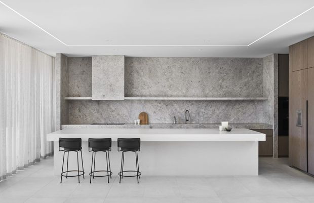 A minimalist contemporary kitchen with a large white island and three black bar stools, set against a grey stone-look backsplash and matching wall cabinetry. The kitchen features a linear integrated rangehood, undermount sink with dark tapware, full-height timber cabinetry at right, pale tiled flooring, sheer floor-to-ceiling curtains, and recessed linear ceiling lighting.