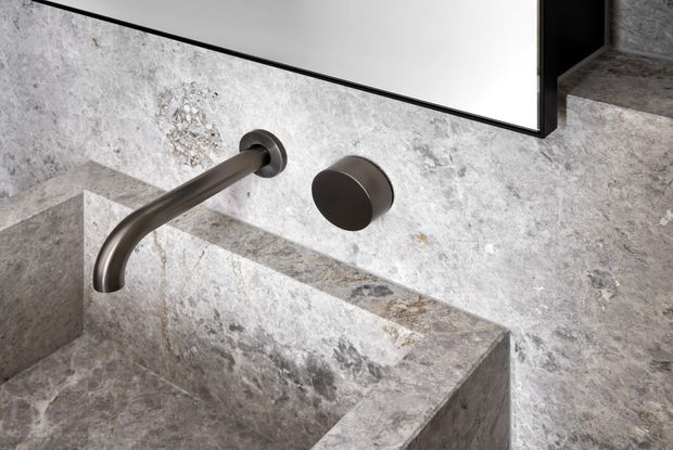 Close-up of a minimalist bathroom basin carved from grey veined natural stone, with a wall-mounted curved dark metal tap and matching round control beneath a frameless mirror. The stone continues across the benchtop, basin, backsplash, and surrounding walls, creating a monolithic refined appearance.