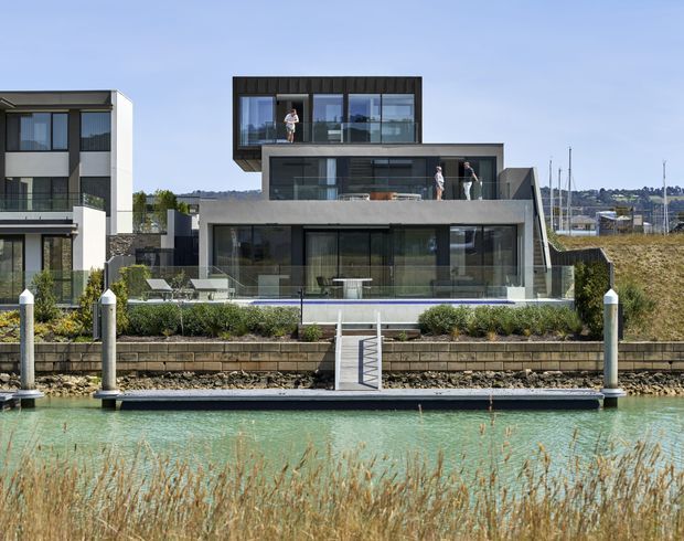 A contemporary multi-level waterfront residence viewed from across a narrow canal. The home features extensive floor-to-ceiling glazing, dark vertical cladding on the upper level, pale rendered or concrete-like walls, broad terraces, glass balustrades, and a private floating dock with steps down to the water. Adjacent modern residences and marina masts are visible in the background.