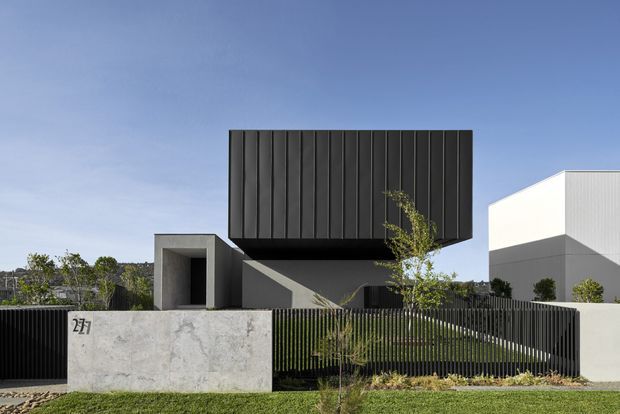 Contemporary two-storey residential facade with a dramatic cantilevered upper volume clad in dark vertical panels above a recessed entry and lower concrete elements. A pale rendered or concrete boundary wall, black vertical-bar fencing, minimalist landscaping, and crisp geometric forms create a high-contrast modern exterior composition.