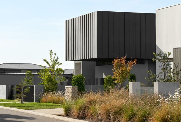 Contemporary multi-level residential facade with a bold cantilevered upper volume clad in dark vertical panels, contrasting with light ribbed cladding and concrete-toned lower walls. The home is framed by a landscaped front garden with ornamental grasses, young trees, low masonry walls, and dark metal fencing.