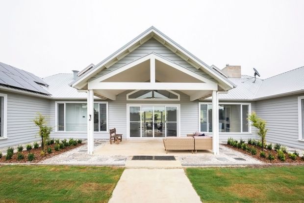 Single-storey residential home exterior with a symmetrical gabled entry porch, white trim, pale grey horizontal weatherboard cladding, and a light metal roof. The covered porch has exposed timber framing and opens onto a straight concrete path, with simple landscaped garden beds and lawn in front. Solar panels are visible on the left roof slope.