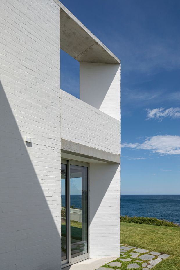 A minimalist white-painted brick coastal house exterior with strong geometric forms, deep rectangular openings, projecting concrete slabs, and a tall glazed door. The building overlooks the ocean, with a manicured lawn and irregular stone stepping path in the foreground under a clear blue sky.