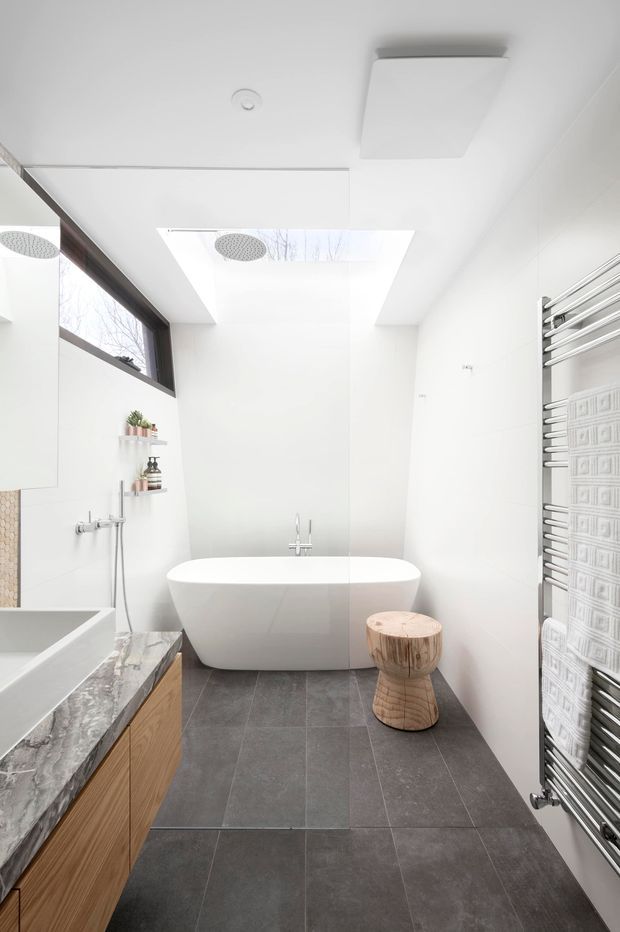 Bright contemporary bathroom with a freestanding white bathtub positioned beneath a large skylight, a glass shower enclosure, and a wall-mounted chrome towel radiator. The room features large dark grey floor tiles, white wall surfaces, a timber vanity with a veined stone benchtop, a partial bathtub at left, and a small sculptural timber stool. A high horizontal window provides additional natural light.