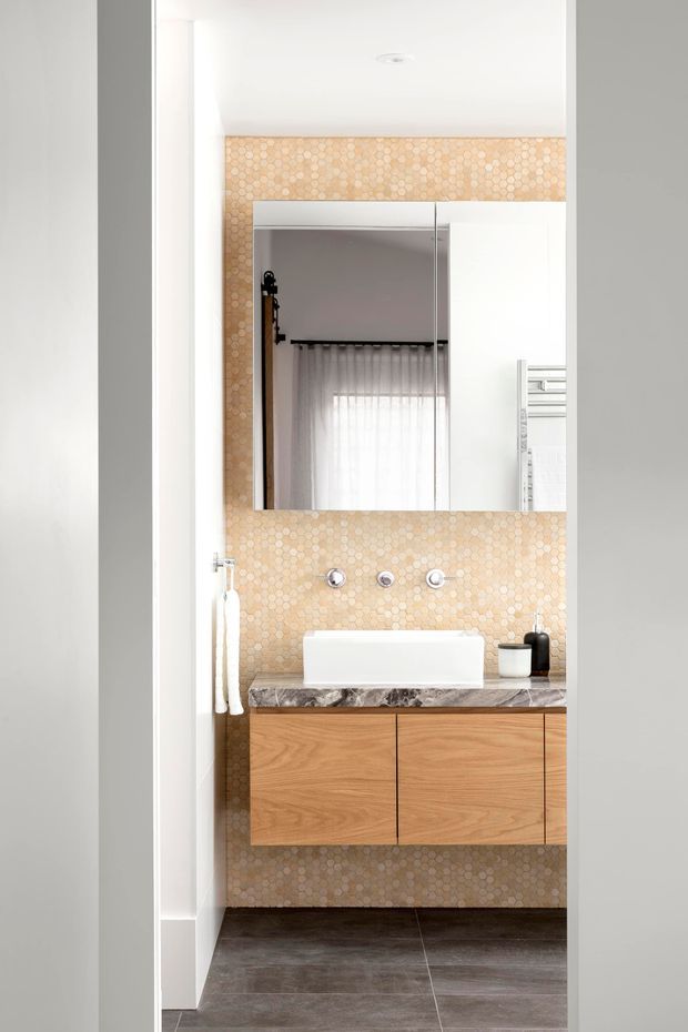 A compact contemporary bathroom viewed through a doorway, featuring a floating timber vanity with a white rectangular basin, dark veined stone benchtop, wall-mounted tapware, and a large frameless mirror. The vanity wall is finished in small warm beige mosaic tiles, while the floor is covered in large dark grey tiles. A heated towel rail is visible at the right edge, and the mirror reflects a softly curtained window.