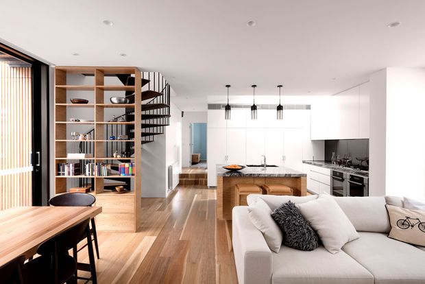 A bright, open-plan contemporary interior combining a kitchen, living room, dining area and circulation space. Warm timber flooring and a timber dining table contrast with white walls and cabinetry. A central kitchen island with integrated timber seating faces a white sectional sofa, while black pendant lights, dark kitchen appliances and a black-framed glazed door add contrast. Open timber shelving forms a partial divider and reveals a sculptural spiral staircase behind it.