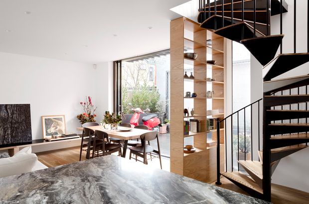 Bright open-plan contemporary interior showing a living and dining area with a dramatic sculptural black spiral staircase, extensive timber open shelving, large glazed openings, and a prominent veined stone kitchen island in the foreground. The room has white walls, timber flooring and joinery, dark dining furniture, and abundant natural light.