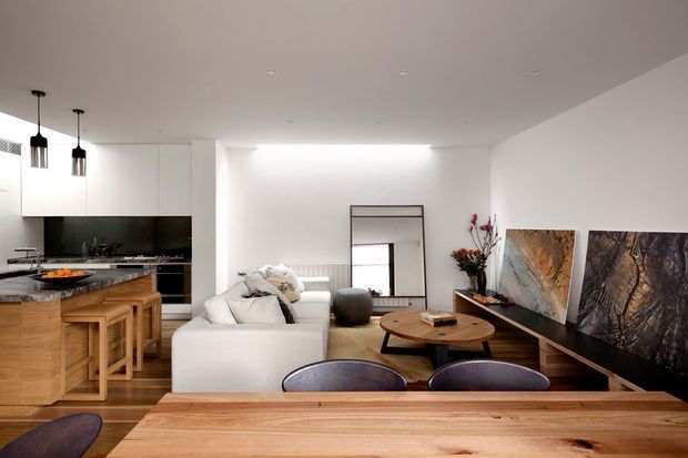 Open-plan contemporary living, dining, and kitchen interior. A white sectional sofa faces a low timber coffee table and large abstract artwork, while a timber dining table occupies the foreground. The kitchen has white handleless cabinetry, a dark splashback, stone benchtops, and timber bar stools. Warm timber flooring and furniture contrast with the predominantly white walls and cabinetry; pendant lights and recessed ceiling lighting provide illumination.