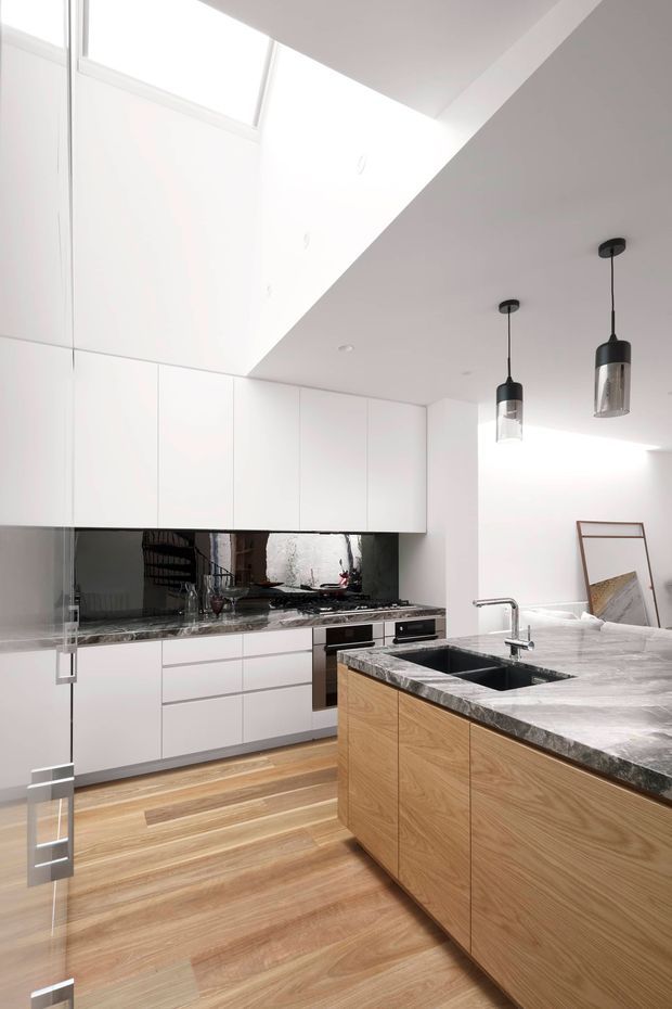 Bright contemporary kitchen with a large timber island, dark stone benchtop, integrated black sink and chrome tapware, white full-height cabinetry, mirrored dark splashback, timber flooring, and black pendant lights. A high ceiling and clerestory-style glazing bring abundant natural light into the space.