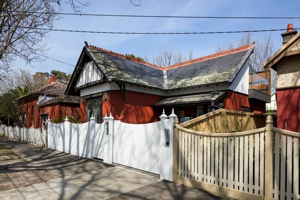 Street-level view of a compact heritage-style residential building with a steeply pitched slate roof, decorative terracotta ridge tiles, half-timbered gable detailing, and deep red painted walls. A white picket-style masonry-post fence and timber gate enclose the front garden, with neighbouring homes and mature deciduous trees along the street.
