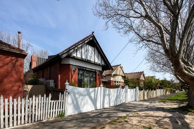 Street-facing view of a compact brick cottage with a prominent front gable, decorative half-timbered infill, dark trim, tiled roof, and a small enclosed front garden. White picket and corrugated-style fencing line the narrow external walkway beneath mature deciduous trees.
