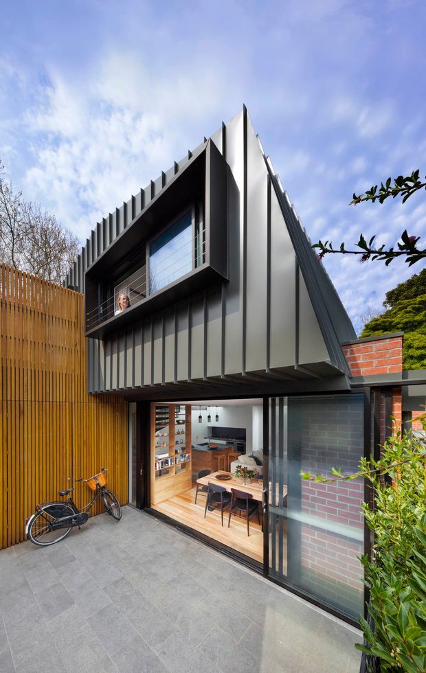 Exterior view of a compact, multi-level contemporary home extension or townhouse. The building features a dramatic steeply angled roof form, dark grey vertical metal cladding with pronounced fins, a projecting upper-level window, exposed red brickwork, and a large glazed opening revealing a warm timber kitchen and dining area. A paved courtyard and timber privacy screen frame the entrance.