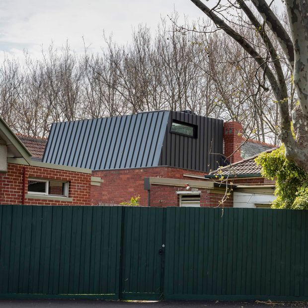 Exterior view of a residential brick house with a contemporary dark corrugated metal rooftop extension, narrow horizontal windows, and a tall dark green privacy fence in the foreground. Mature deciduous trees surround the property, and the extension contrasts with the existing low-pitched tiled roof and red brick walls.