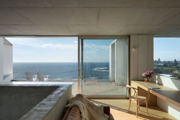 A bright contemporary interior with floor-to-ceiling glazing opening onto a balcony and expansive ocean views. The room includes a concrete-clad built-in feature or partition in the foreground, warm timber flooring, a minimalist built-in desk or console, and a timber-framed chair. An adjacent coastal residential setting is visible through the windows.