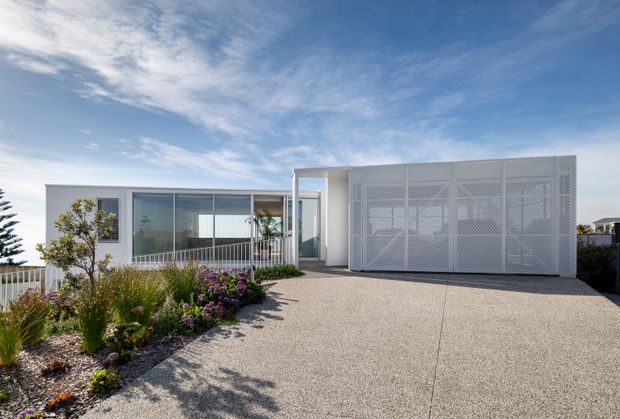 Front exterior view of a contemporary single-storey home with extensive floor-to-ceiling glazing, a white rendered facade, and a prominent perforated white screen enclosing the garage or carport. A broad textured concrete driveway leads to the entry, with low landscaped planting along the edge.