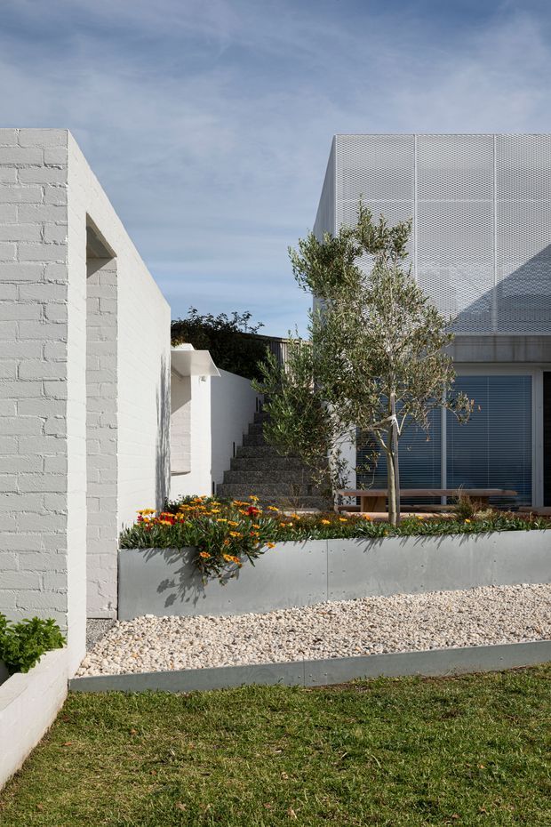 Exterior view of a contemporary residential property with white-painted masonry walls, geometric volumes, a perforated metal upper facade, dark-framed glazing, and landscaped stepped garden beds. A gravel forecourt, planted retaining wall, mature tree, and exterior stairway are visible under a bright blue sky.