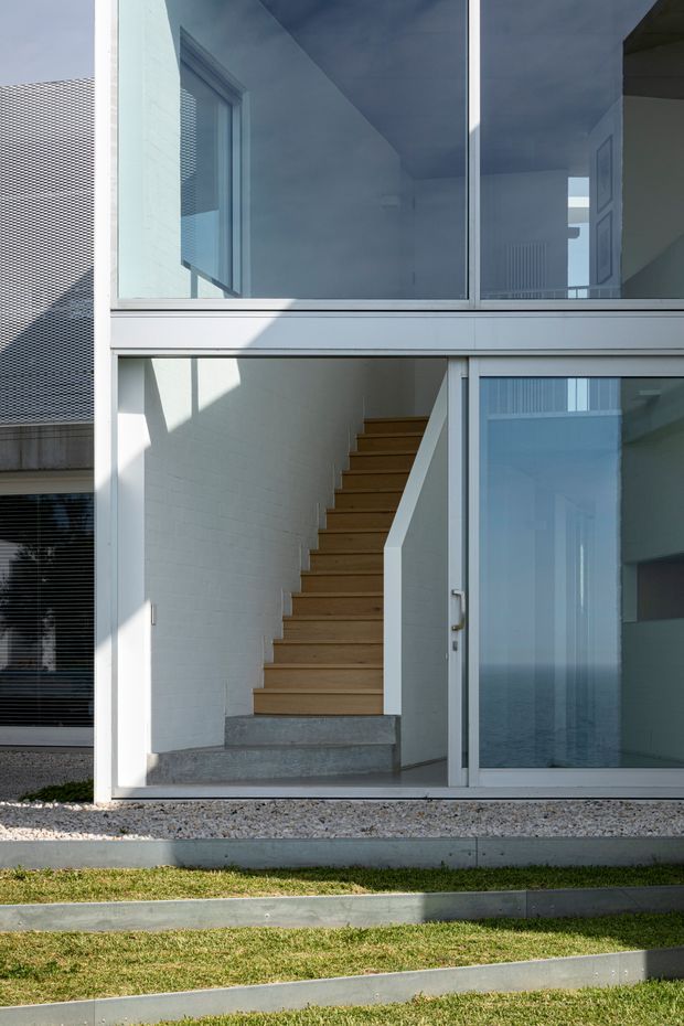 Exterior architectural view of a contemporary multi-level dwelling, focused on a glazed stairwell and entry enclosure. Large blue-tinted windows and sliding glass doors are set in pale metal frames, revealing a straight timber staircase against white painted masonry walls. Concrete steps and edging sit in a tiered lawn and gravel foreground, with strong sunlight and shadows defining the minimalist facade.