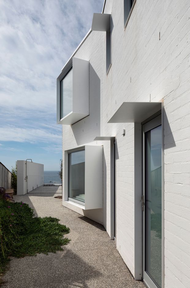 A narrow exterior side passage alongside a contemporary white-painted brick house. The facade features projecting box-like window surrounds and deep rectangular sun-shading hoods around dark-framed glazed openings. A textured light aggregate path runs beside low planting toward a gated terrace, with a glimpse of blue water or horizon beyond. The image has a clean, minimalist composition under a bright partly cloudy sky.