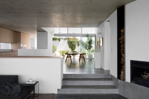 Open-plan contemporary interior showing a kitchen at left, a dining area beyond, and a living area at right with a built-in fireplace and stacked firewood niche. The space has a raw concrete ceiling, polished grey concrete floors and steps, white walls, light timber kitchen cabinetry, and large glazed openings to a planted courtyard or garden. The room is bright and minimalist with strong indoor-outdoor connection.