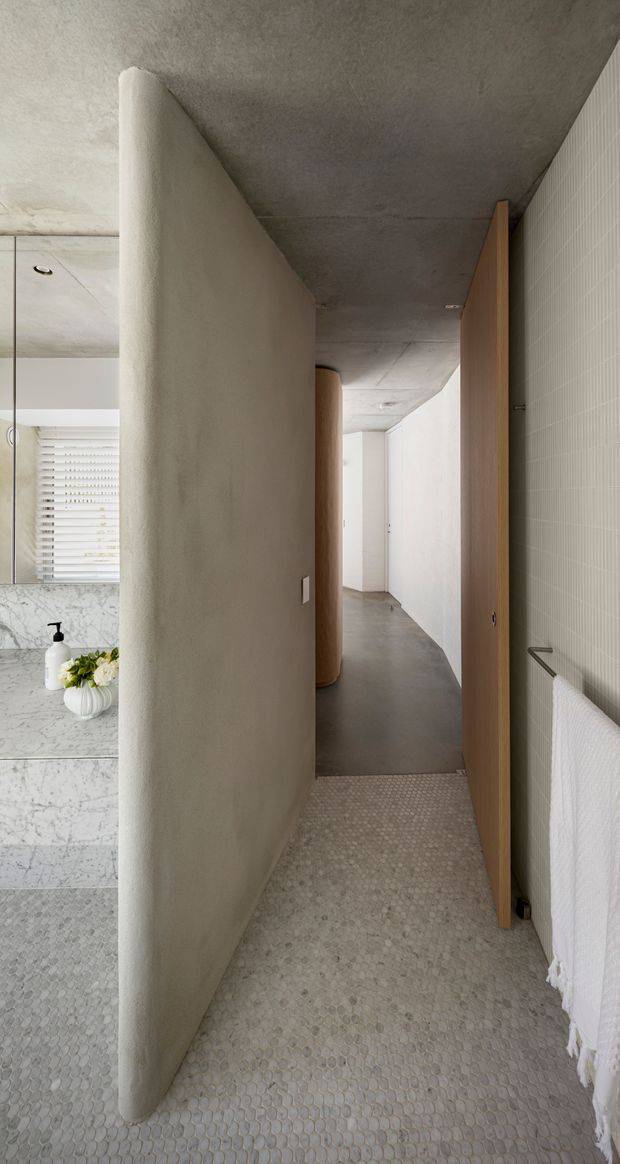 A narrow, refined bathroom passage with a curved plaster privacy wall dividing the vanity area from a hallway and enclosed wet-room spaces. The bathroom features pale small-format mosaic floor tiles, a marble vanity and backsplash, concrete-look ceiling and corridor floor, warm timber doors, white walls, and a minimalist towel rail. A large mirror and window blinds are visible beside the vanity, creating a light, textured contemporary interior.