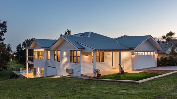 Exterior view of a large, light-coloured weatherboard house at dusk, featuring multiple pitched gable roofs, extensive windows, an attached garage, a covered entry porch, and a rear balcony or terrace overlooking a sloping landscaped lawn.