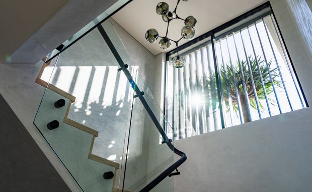 Bright contemporary stairwell with a sculptural dark handrail, clear glass balustrade, pale textured walls, and a large corner window fitted with vertical black louvres. Sunlight casts strong linear shadows across the stair enclosure, while a clustered chrome-and-glass pendant light hangs overhead. Tropical foliage is visible outside the glazing.