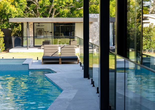 View across a modern pool area toward a low-slung pavilion or house, with a turquoise swimming pool, pale paved pool surround, streamlined dark-framed floor-to-ceiling glazing, and a covered outdoor lounging area. Mature trees and landscaped greenery surround the property. The composition highlights the relationship between the pool, outdoor seating, and glazed contemporary architecture.