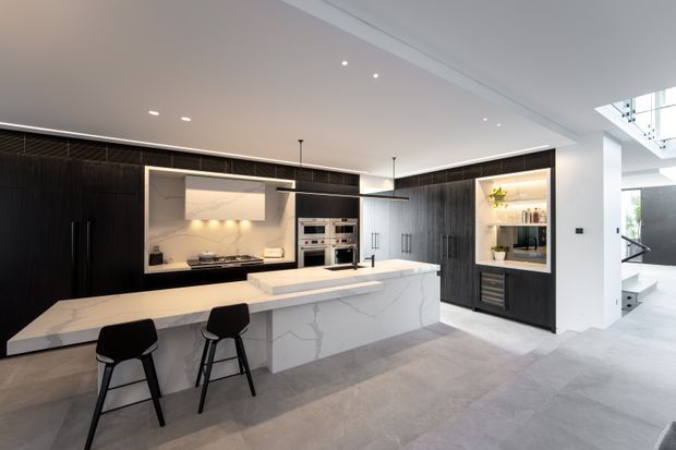 A spacious contemporary kitchen with a large white marble-look island and integrated breakfast bar, dark full-height cabinetry, concealed rangehood, built-in ovens, and open display shelving. Pale large-format flooring, recessed ceiling lighting, and linear pendant lights create a clean, high-contrast interior. A bright stairwell or adjoining circulation area is visible at the right.