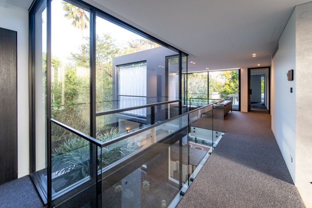 A bright contemporary upper-level hallway or landing overlooking a planted internal courtyard through extensive floor-to-ceiling glazing. The space has a dark carpeted floor, white walls and ceiling, slim black-framed windows, and a frameless glass balustrade around an open void and stairwell. A corridor leads toward additional glazed rooms and exterior views.